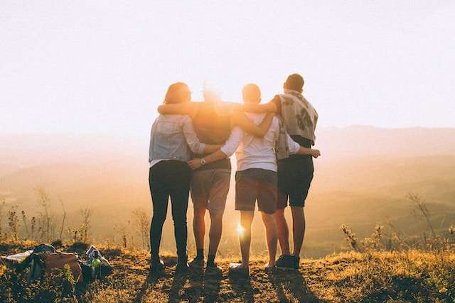 Keturi draugai apsikabinę žiūri į saulėlydį kalnuose, simbolizuojantys draugystę.