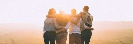 Keturi draugai apsikabinę žiūri į saulėlydį kalnuose.